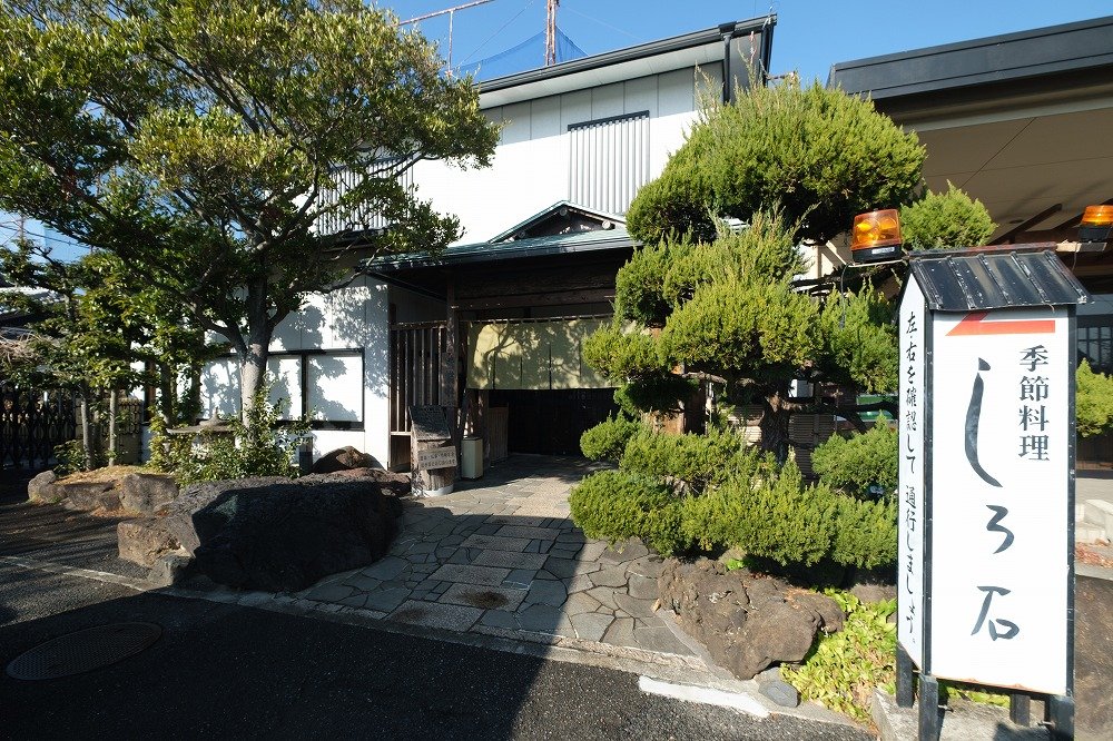 植栽のある門まわりと落ち着いた建物外観、その前に駐車スペースが広がる店舗全景